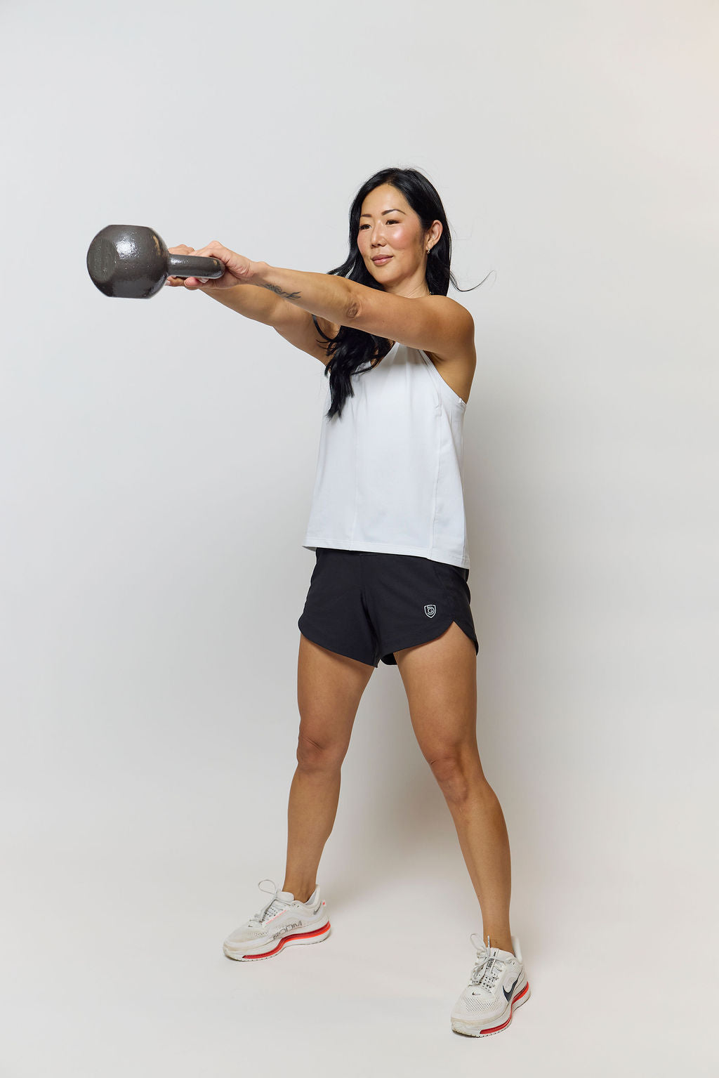 A woman in athletic wear holding a dumbbell overhead in a studio setting. She is wearing a white sleeveless top, black athletic shorts, and white sneakers with red accents.