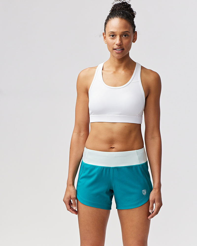 Woman wearing a white sports bra and teal shorts on a light gray background
