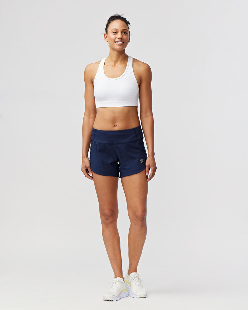 Woman wearing a white sports bra and navy shorts on a light gray background