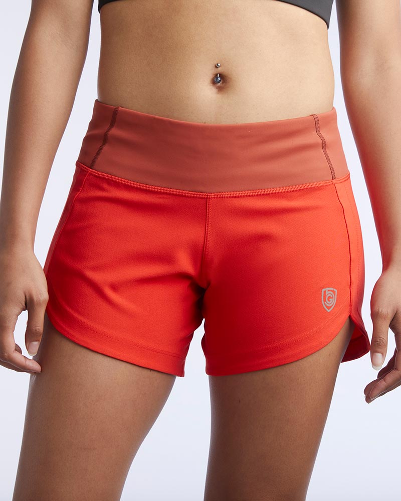 A close-up of a model wearing bright red athletic shorts with a wide, slightly sheer orange-brown waistband. The shorts feature a small shield logo on the lower left leg and are shown against a plain light gray background.