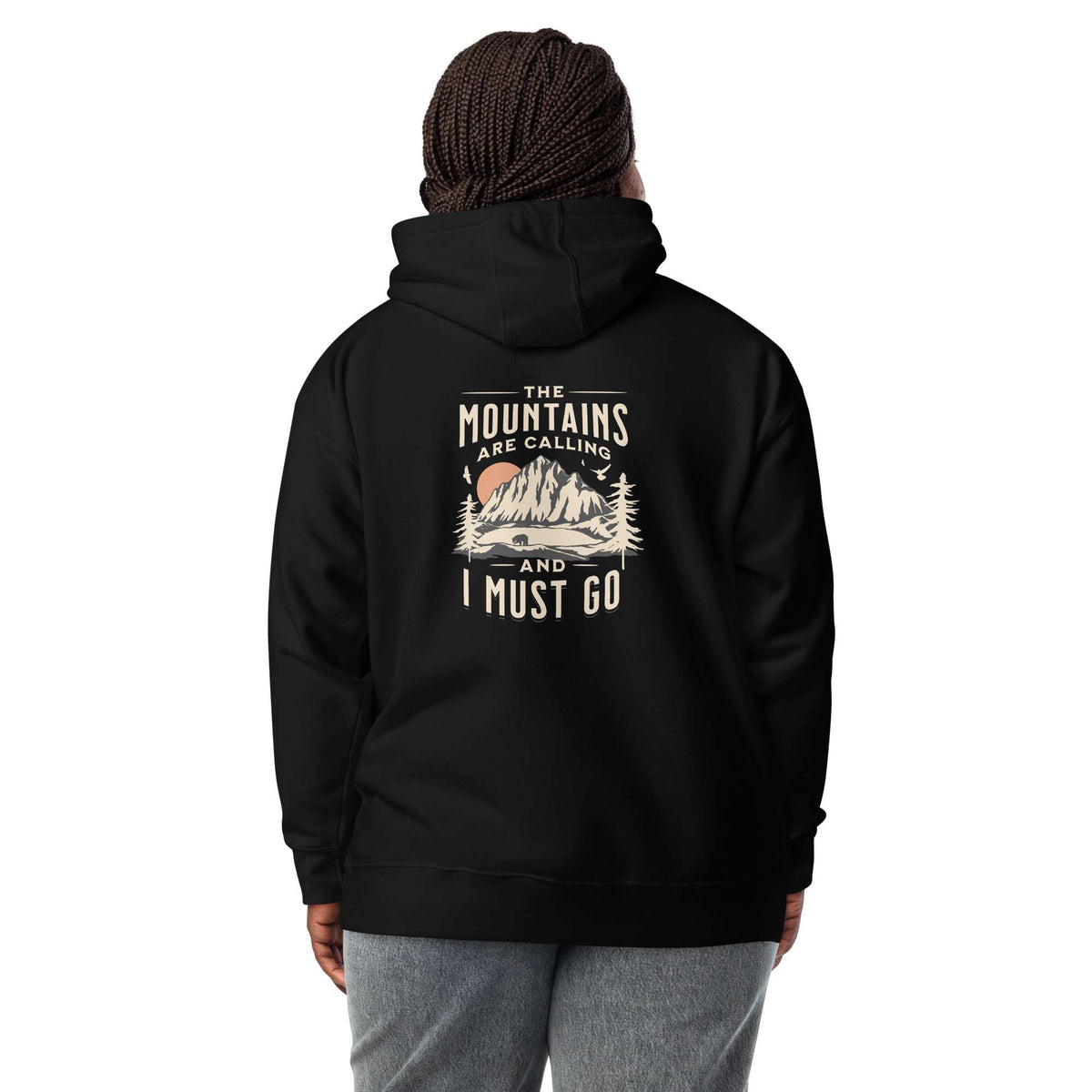 A person shown from the back wearing a black pullover hoodie with a vintage-style mountain graphic and the text 'THE MOUNTAINS ARE CALLING AND I MUST GO' printed on the back. The person is also wearing a dark knit beanie and gray sweatpants against a plain white studio background. Nature Quote Mountains Are Calling Graphic Hoodie Casual Wear Shop the Nature Quote Mountains Are Calling Graphic Hoodie, a cozy statement piece for hikers, campers, and everyday adventure seekers.