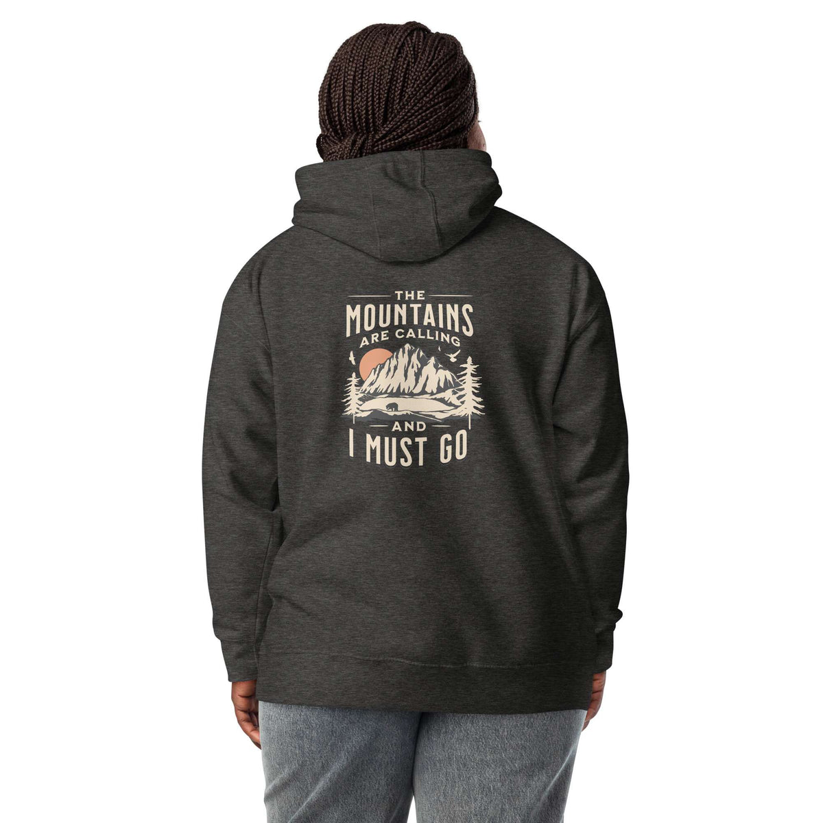 A person shown from the back wearing a dark heather charcoal hoodie with a vintage-style mountain graphic and the text 'THE MOUNTAINS ARE CALLING AND I MUST GO' printed on the back. The person is also wearing gray sweatpants and a dark brown beaded hat, set against a plain white background. Nature Quote Mountains Are Calling Graphic Hoodie Casual Wear Shop the Nature Quote Mountains Are Calling Graphic Hoodie, a cozy statement piece for hikers, campers, and everyday adventure seekers.
