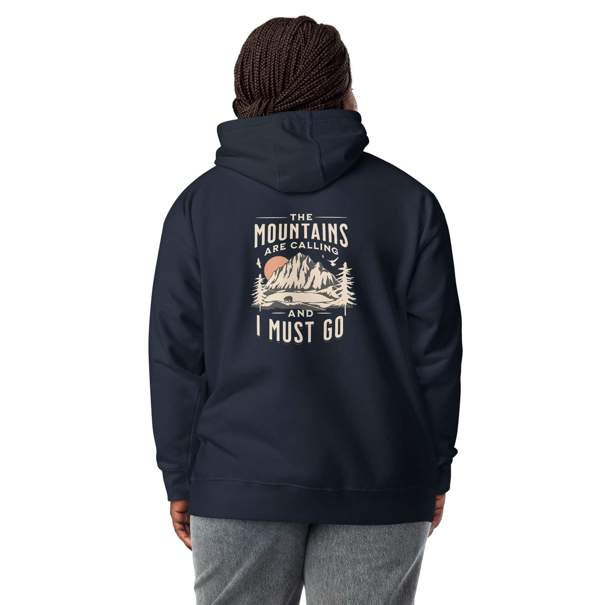 A person shown from the back wearing a navy pullover hoodie with a large vintage-style mountain graphic and the text 'THE MOUNTAINS ARE CALLING AND I MUST GO' printed on the back. The model is also wearing gray sweatpants and a dark knit beanie, photographed against a plain white studio background.