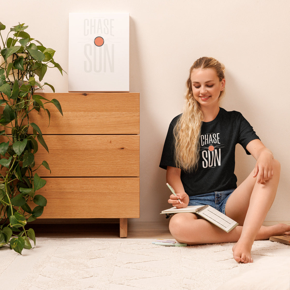 A woman sits on a light-colored floor next to a wooden dresser, wearing a black graphic t-shirt with 'CHASE SUN' printed on the front. A matching framed print hangs on the dresser above her, and a green houseplant is on the left side of the scene. Chase the Sun Minimalist Lettering Graphic T-Shirt Casual Wear Make every day feel brighter in the Chase the Sun Minimalist Lettering Graphic T-Shirt, a simple statement tee with relaxed style.